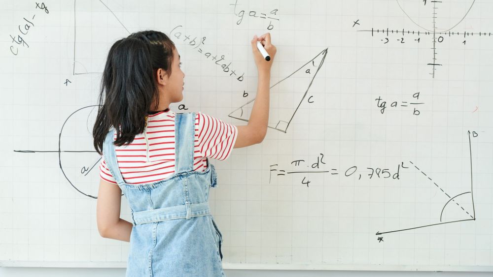 Smart schoolgirl writing on whiteboard when solving geometry task in class, view from the back